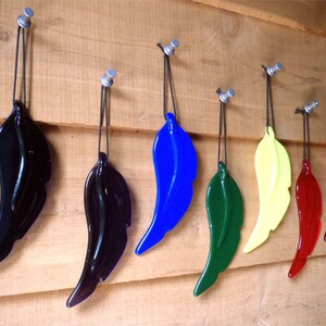 Puede incluir: Siete adornos de plumas de vidrio colgando de ganchos en una pared de madera. Las plumas son de varios colores, incluyendo negro, azul, verde, amarillo y rojo.