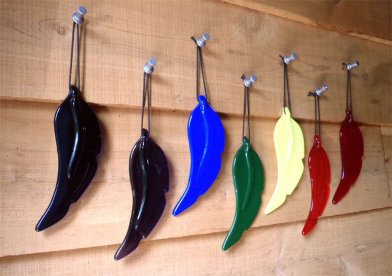 Puede incluir: Siete adornos de plumas de vidrio colgando de ganchos en una pared de madera. Las plumas son de varios colores, incluyendo negro, azul, verde, amarillo y rojo.
