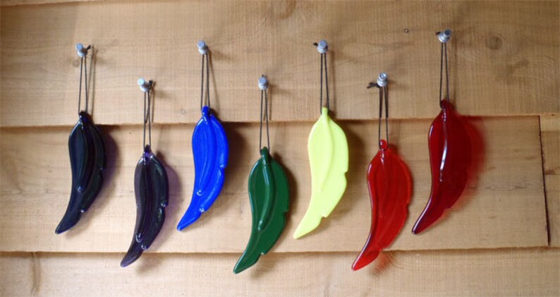 Puede incluir: Siete adornos de plumas de vidrio colgando de ganchos en una pared de madera. Las plumas son de varios colores, incluyendo azul, verde, amarillo, negro y rojo.
