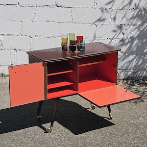 Può includere: Un carrello bar in legno con piano marrone scuro e interno color corallo brillante. È dotato di un'anta a battente, un'anta a ribalta e quattro gambe affusolate con rotelle dorate. Un portabicchieri in metallo con cinque bicchieri colorati è appoggiato sopra.