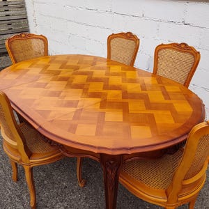 May include: An oval wooden dining table with a light and dark brown chevron parquet pattern on the surface, featuring four matching chairs with woven cane backrests and wooden frames, all displaying a polished finish and carved detailing.