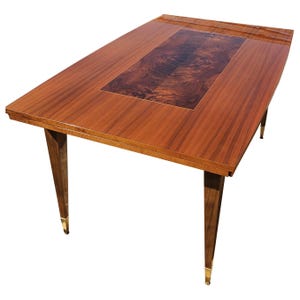 Peut inclure: Une table à manger rectangulaire avec une surface en bois poli présentant une incrustation centrale en ronce de bois foncé. La table possède quatre pieds fuselés avec des embouts dorés métalliques à la base et une finition brillante.