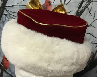 Può includere: Un cappello festivo con corona in velluto rosso e finiture dorate, impreziosito da due ornamenti dorati a forma di fiocco. Il cappello presenta un'ampia fascia di soffice pelliccia sintetica bianca. Il cappello è progettato per le festività.
