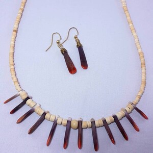 May include: A necklace and earrings set made with white and red beads and red and purple shell pieces. The necklace has a graduated color pattern of red, purple, white, and beige beads. The earrings are simple gold hoops with a red and purple shell piece hanging from each.