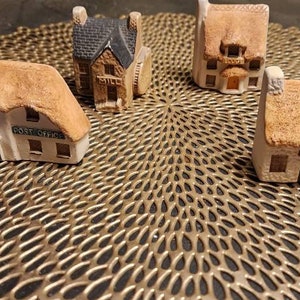 May include: Four miniature houses with thatched roofs, painted white with brown accents, are arranged on a gold circular placemat. The houses are decorated with details like windows, doors, and chimneys. One house has the words "Post Office" written on the front.