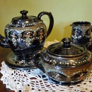 May include: A black tea set with gold and silver accents. The teapot, creamer, and sugar bowl are decorated with floral patterns.