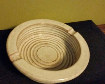 May include: White ceramic ashtray with a circular, concentric ring design. The ashtray has a wide, shallow bowl.
