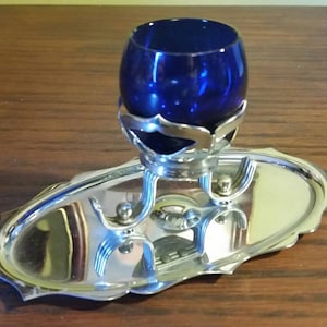 May include: A silver-colored, oval-shaped serving tray with a decorative rim. A cobalt blue glass sits in a silver holder above the tray. The tray has a decorative design with two small silver accents.