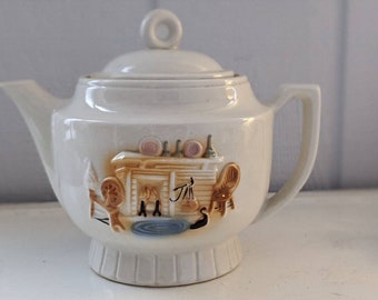 May include: A white ceramic teapot with a brown and tan scene of a fireplace and chairs. The teapot has a lid with a knob on top.