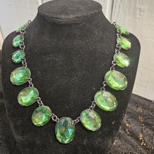 May include: A green gemstone necklace with oval-shaped stones. The stones are linked together with a dark metal chain. The necklace is displayed on a black mannequin.