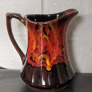 May include: A small, brown ceramic pitcher with a red, yellow, and black glaze. The pitcher has a flared spout and a handle.
