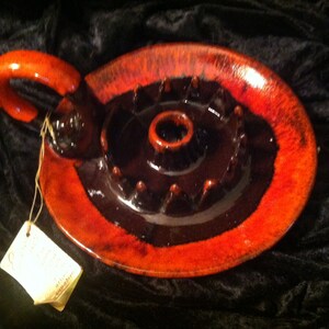 May include: A red and black ceramic candle holder with a unique design. The holder has a circular base with a raised center and a handle on the side.
