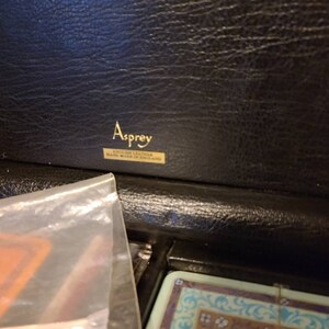 Op de afbeelding: Een zwarte leren doos met een gouden label met de tekst "Asprey English Leather Hand Made in England."