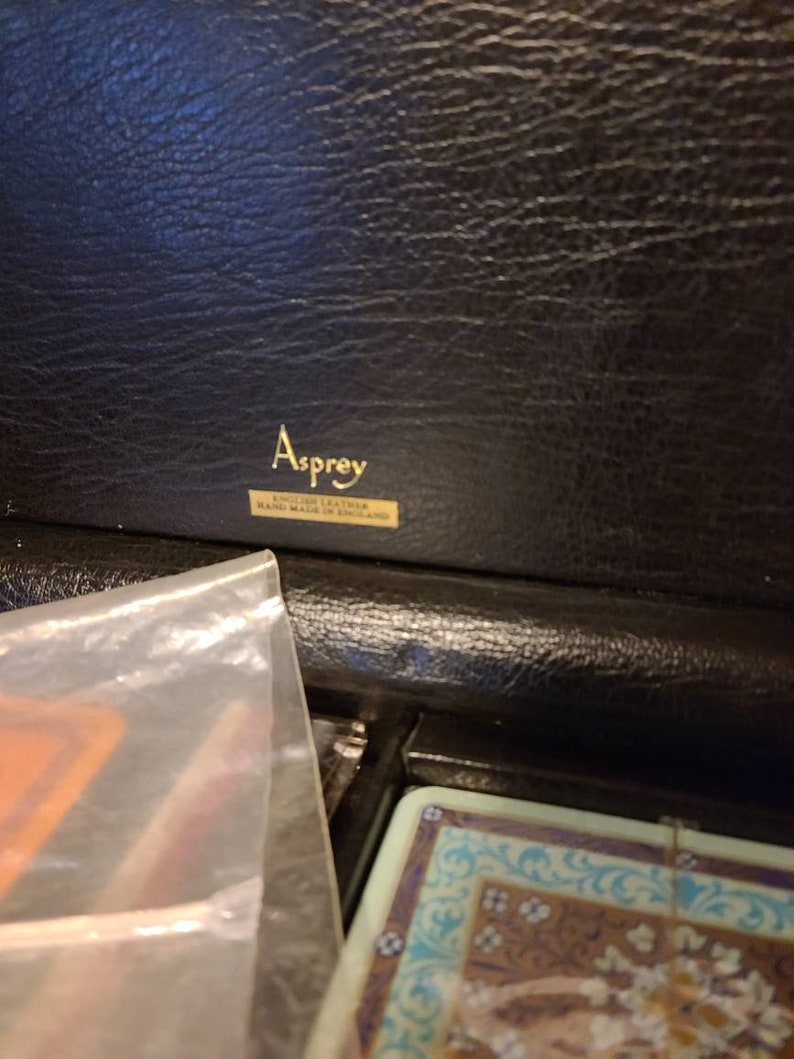 Op de afbeelding: Een zwarte leren doos met een gouden label met de tekst "Asprey English Leather Hand Made in England."