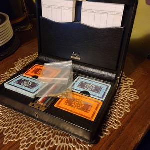 Op de afbeelding: Een zwart leren kaartenetui met een gouden slot en sleutel. De koffer is open en bevat twee kaartspellen, een met een blauw-wit ontwerp en een met een oranje-bruin ontwerp. Er zijn ook twee potloden en een klein plastic zakje.