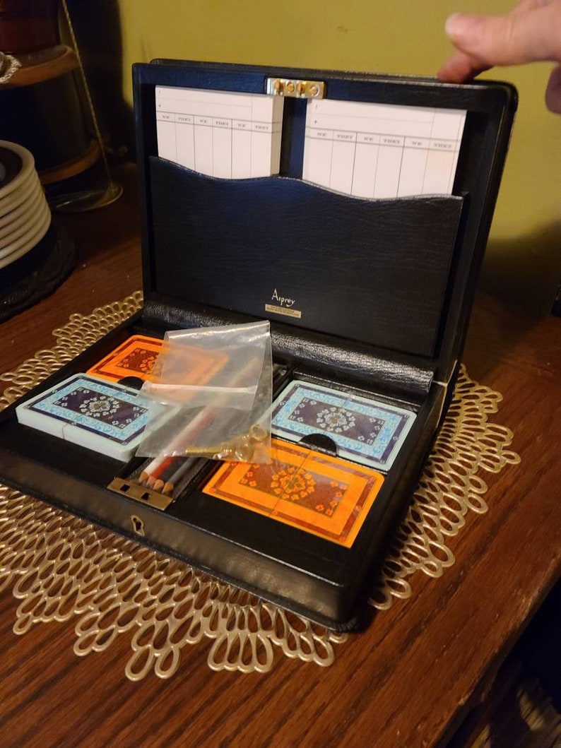 Op de afbeelding: Een zwart leren kaartenetui met een gouden slot en sleutel. De koffer is open en bevat twee kaartspellen, een met een blauw-wit ontwerp en een met een oranje-bruin ontwerp. Er zijn ook twee potloden en een klein plastic zakje.
