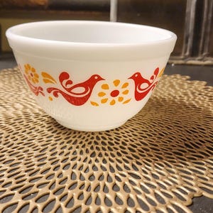 May include: White mixing bowl with a red and yellow bird design. The bowl is sitting on a gold-colored doily.