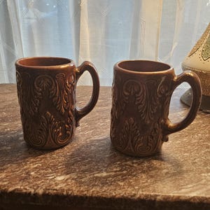 May include: Two brown ceramic mugs with ornate, raised floral patterns. Each mug has a curved handle and a dark brown interior. The mugs are displayed on a brown surface.