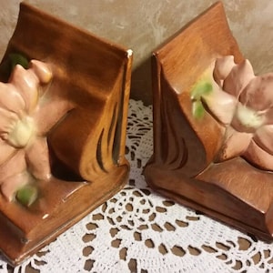 May include: A pair of bookends made of wood and ceramic. Each bookend features a pink flower with green leaves. The bookends are a dark brown color.