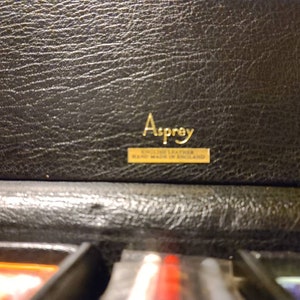 Op de afbeelding: Een zwarte leren doos met een gouden label waarop staat "Asprey, English Leather, Made in England."