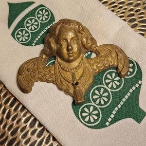 May include: A gold-toned angel ornament with wings and a cherub face. The ornament is on a white fabric with a green and white design.