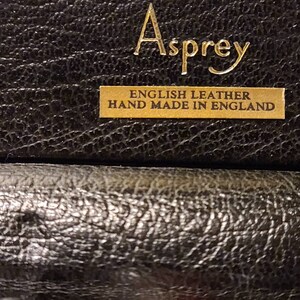 Op de afbeelding: Close-up van een zwarte leren doos met gouden letters die "Asprey English Leather Hand Made in England" lezen.