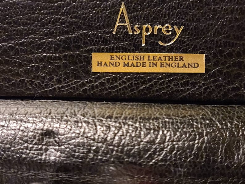 Op de afbeelding: Close-up van een zwarte leren doos met gouden letters die "Asprey English Leather Hand Made in England" lezen.