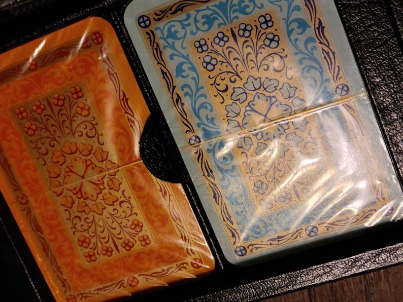 Op de afbeelding: Twee speelkaarten met een bloemenmotief. De kaart links is oranje en goud met een rood bloemenpatroon. De kaart rechts is blauw en goud met een groen bloemenpatroon.