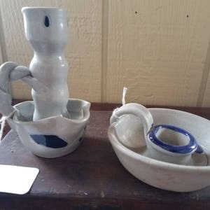 May include: Two ceramic candle holders with blue and white glaze. One holder is a tall, twisted design with a wide base. The other holder is a bowl shape with a handle and a small blue rim.
