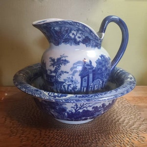 May include: A blue and white ceramic pitcher and bowl set. The pitcher has a curved handle and spout, and the bowl is wide and shallow. Both pieces feature a detailed landscape design. The set is placed on a gold-colored decorative mat.