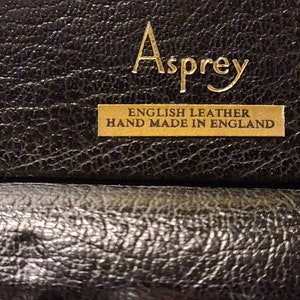Op de afbeelding: Close-up van een zwarte leren doos met gouden letters die "Asprey English Leather Hand Made in England" lezen.