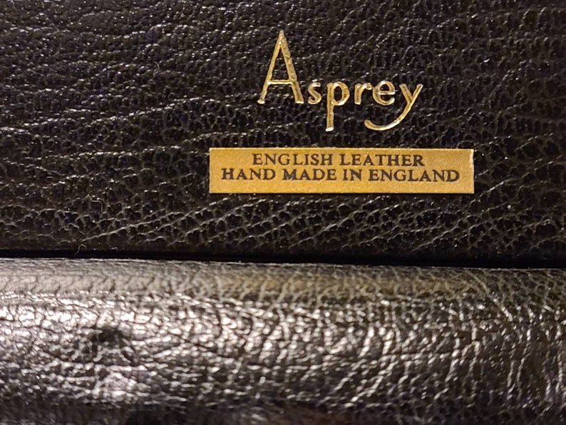 Op de afbeelding: Close-up van een zwarte leren doos met gouden letters die "Asprey English Leather Hand Made in England" lezen.