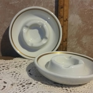 May include: Two white ceramic ashtrays with a gold rim. The ashtrays are round and have a shallow well in the center.