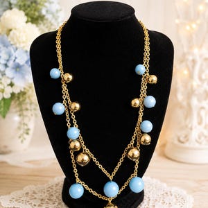 May include: A multi-strand gold-tone chain necklace featuring alternating light blue spherical beads and polished gold-tone metallic spheres. The necklace is displayed on a black bust, showcasing a layered design with dangling beads throughout the length.