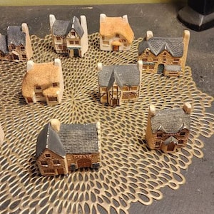 May include: A collection of miniature ceramic houses in various styles and colors, arranged on a gold-colored doily. The houses are all different sizes and shapes, and some have windows and doors.