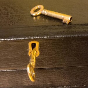 Op de afbeelding: Een gouden sleutel met een rond sleutelgat ligt op een zwarte lederen doos. De sleutel is gedeeltelijk in het sleutelgat van de doos gestoken.
