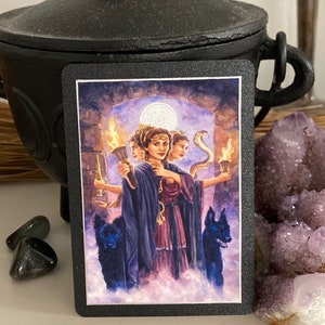 K&ouml;nnte beinhalten: Tarotkarte mit einer Frau mit drei Gesichtern, die jeweils eine Fackel halten. Sie ist von zwei schwarzen Hunden und einer Schlange umgeben. Die Karte ist vor dem Hintergrund eines Steinbogens und eines bew&ouml;lkten Himmels platziert.