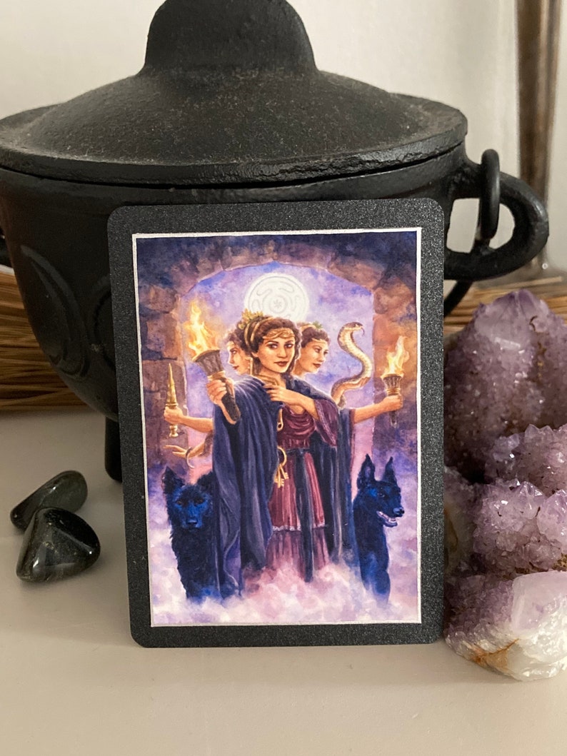 K&ouml;nnte beinhalten: Tarotkarte mit einer Frau mit drei Gesichtern, die jeweils eine Fackel halten. Sie ist von zwei schwarzen Hunden und einer Schlange umgeben. Die Karte ist vor dem Hintergrund eines Steinbogens und eines bew&ouml;lkten Himmels platziert.