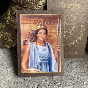 May include: A tarot card featuring a woman in a blue dress, holding a spear and shield, with an owl on her shoulder. The card has a brown border and the Greek word "Athena" is written on the card. Another card with an owl illustration is in the background.