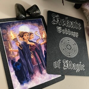 K&ouml;nnte beinhalten: Eine schwarze Karte mit silbernem Text und einem silbernen Symbol. Der Text lautet "Hekate G&ouml;ttin der Magie". Das Symbol ist ein Kreis mit einem Stern in der Mitte und vier konzentrischen Kreisen darum herum.