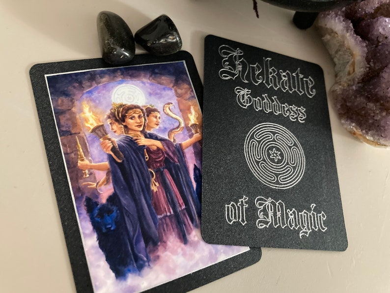 K&ouml;nnte beinhalten: Eine schwarze Karte mit silbernem Text und einem silbernen Symbol. Der Text lautet "Hekate G&ouml;ttin der Magie". Das Symbol ist ein Kreis mit einem Stern in der Mitte und vier konzentrischen Kreisen darum herum.