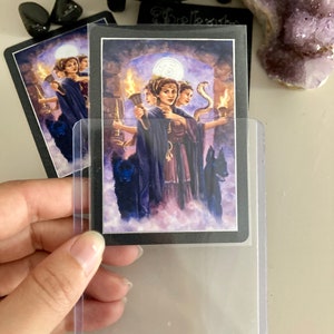 K&ouml;nnte beinhalten: Eine Tarotkarte mit schwarzem Hintergrund und einer lila und wei&szlig;en Illustration von drei Frauen mit einer Schlange und einem Vollmond im Hintergrund. Die Karte befindet sich in einer durchsichtigen Plastikh&uuml;lle.