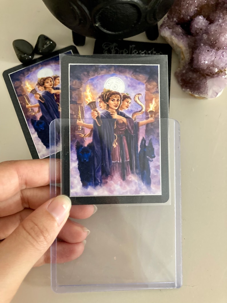 K&ouml;nnte beinhalten: Eine Tarotkarte mit schwarzem Hintergrund und einer lila und wei&szlig;en Illustration von drei Frauen mit einer Schlange und einem Vollmond im Hintergrund. Die Karte befindet sich in einer durchsichtigen Plastikh&uuml;lle.