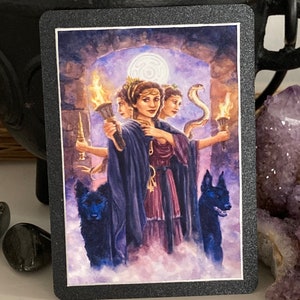 K&ouml;nnte beinhalten: Eine Tarotkarte mit einem dunklen Hintergrund und einer Frau mit drei Gesichtern, die eine Fackel h&auml;lt. Die Frau tr&auml;gt eine violette Robe und eine Krone. Zwei schwarze Hunde stehen zu beiden Seiten von ihr.