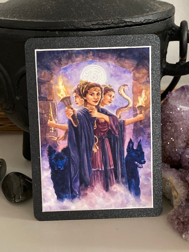 K&ouml;nnte beinhalten: Eine Tarotkarte mit einem dunklen Hintergrund und einer Frau mit drei Gesichtern, die eine Fackel h&auml;lt. Die Frau tr&auml;gt eine violette Robe und eine Krone. Zwei schwarze Hunde stehen zu beiden Seiten von ihr.