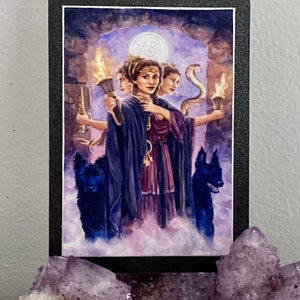 K&ouml;nnte beinhalten: Tarotkarten-Illustration, die eine Frau mit drei K&ouml;pfen zeigt, jeder mit einem anderen Ausdruck. Sie tr&auml;gt eine lila Robe und h&auml;lt eine Fackel. Zwei schwarze Hunde stehen zu beiden Seiten von ihr. Der Hintergrund ist eine dunkle, geheimnisvolle Szene mit einem Mond am Himmel.