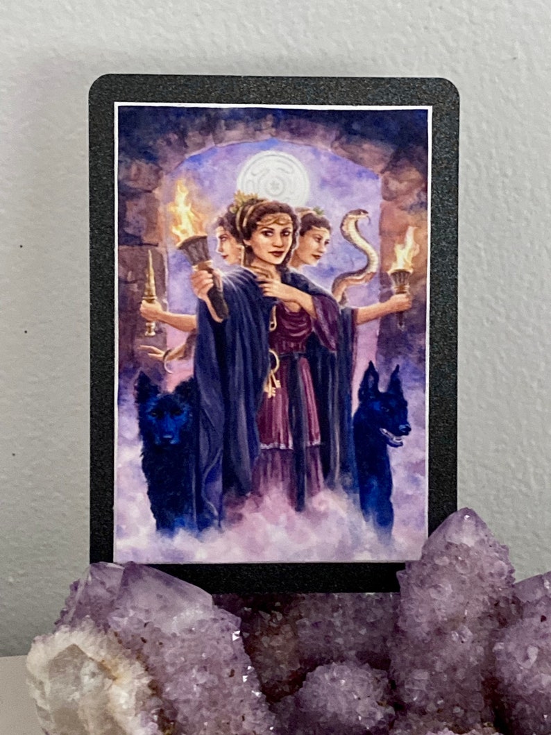 K&ouml;nnte beinhalten: Tarotkarten-Illustration, die eine Frau mit drei K&ouml;pfen zeigt, jeder mit einem anderen Ausdruck. Sie tr&auml;gt eine lila Robe und h&auml;lt eine Fackel. Zwei schwarze Hunde stehen zu beiden Seiten von ihr. Der Hintergrund ist eine dunkle, geheimnisvolle Szene mit einem Mond am Himmel.