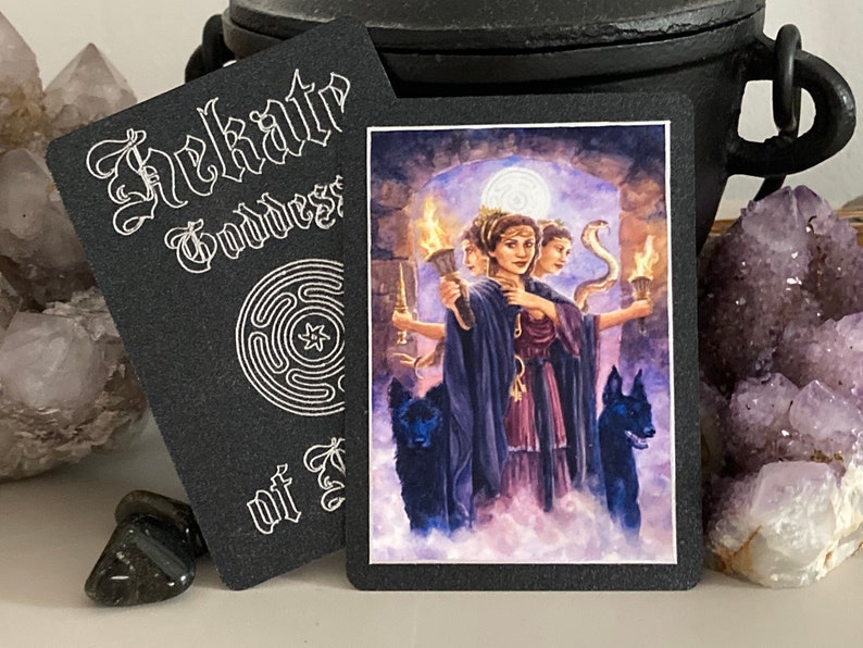 K&ouml;nnte beinhalten: Eine schwarze Karte mit wei&szlig;em Text, der "Hekate G&ouml;ttin der Kreuzwege" lautet, und einem wei&szlig;en Labyrinth-Design. Die Karte zeigt ein farbenfrohes Bild einer dreik&ouml;pfigen G&ouml;ttin mit Schlangen und Fackeln.