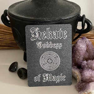 K&ouml;nnte beinhalten: Eine schwarze Karte mit wei&szlig;em Text, der "Hekate G&ouml;ttin der Magie" lautet. Ein wei&szlig;es Symbol mit einem Stern in der Mitte befindet sich auf der Karte. Die Karte liegt auf einer wei&szlig;en Oberfl&auml;che mit einem schwarzen Kessel und violetten Kristallen.