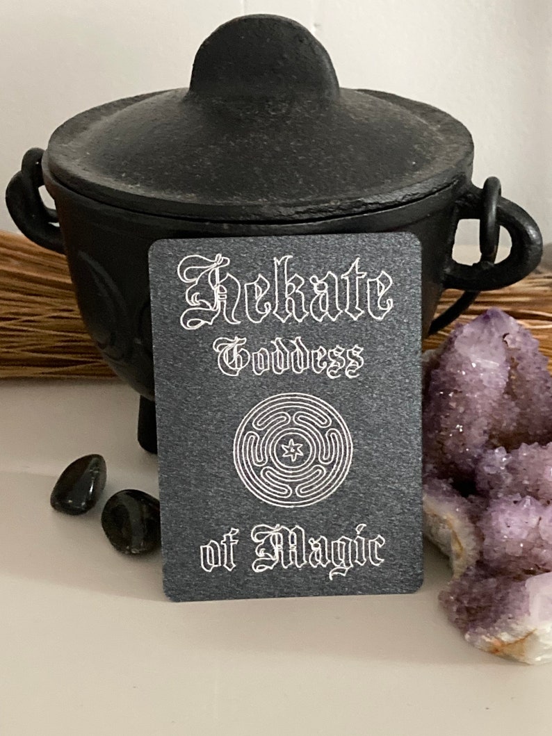 K&ouml;nnte beinhalten: Eine schwarze Karte mit wei&szlig;em Text, der "Hekate G&ouml;ttin der Magie" lautet. Ein wei&szlig;es Symbol mit einem Stern in der Mitte befindet sich auf der Karte. Die Karte liegt auf einer wei&szlig;en Oberfl&auml;che mit einem schwarzen Kessel und violetten Kristallen.
